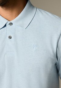 Vaaleansininen polo-paita, joka on valmistettu teksturoidusta kankaasta, jossa on kaulus, kaksi harmaata nappia ja pieni brodeerattu hirvilogot rinnassa.