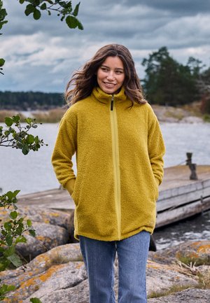 Jeune femme en veste polaire jaune et jean debout sur une rive rocheuse près d'un quai en bois sous un ciel nuageux.
