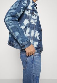 Homme en veste en denim bleu délavé avec texte blanc et jean assorti tenant l'ourlet de la veste avec la main gauche.