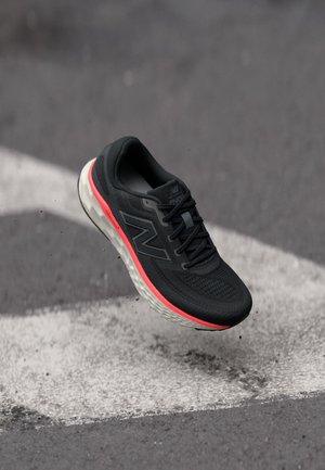 Chaussure de course New Balance noire et grise avec un détail de semelle rouge flottant au-dessus d’un bitume avec des lignes blanches peintes.