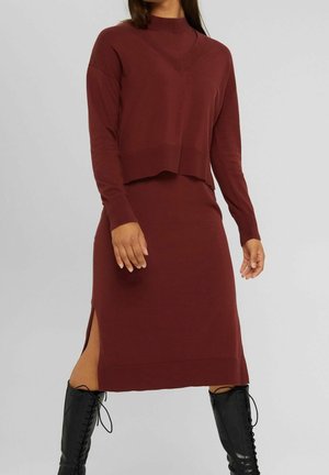 Vínová pletená dvoudílná souprava s krátkým topem s nákrčníkem a midi sukní s bočním rozparkem, doplněná černými šněrovacími kozačkami nad kolena.