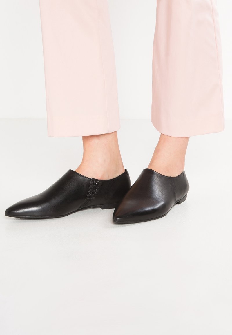 Paire de chaussures en cuir noir à bout pointu, sans lacets, portées avec un pantalon court rose clair sur fond clair.