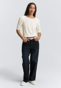 Witte top met korte mouwen en pofmouwen, gecombineerd met donkerblauwe brede jeans en een beige riem. Witte sneakers maken de look compleet.