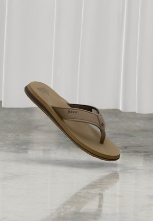 Tanfarbene Flip-Flop-Sandale mit strukturiertem Riemen und "REEF"-Logo, schwebend über einem reflektierenden Marmorboden vor weißem Vorhanghintergrund.