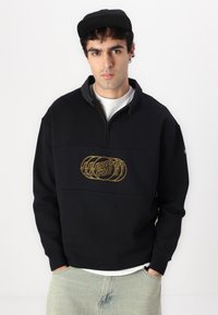 Sudadera negra con cremallera en el cuello y un cuello gris, con un diseño de logo dorado en el pecho. Material suave y texturizado, con un ajuste relajado.