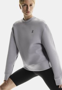 Sweatshirt lilas clair avec un col rond, des poignets côtelés et un logo subtil sur la poitrine, associé à un short de vélo noir. Texture lisse.