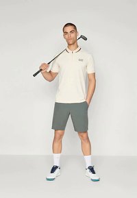 Πόλο πουκάμισο γκολφ σε off-white απόχρωση με κόκκινες λεπτομέρειες στον γιακά, συνδυασμένο με γκρι σορτς. Το μοντέλο κρατά μια γκολφ κλαμπ, φορώντας λευκά παπούτσια με μπλε λεπτομέρειες.