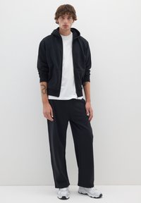 Schwarze Kapuzenjacke mit Reißverschluss in lockerer Passform, kombiniert mit schwarzen Hosen. Das Outfit besteht aus einem weißen T-Shirt und grauen Sneakers.