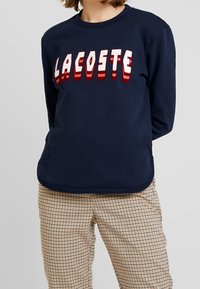 Marinblå crewneck sweatshirt i bomullsmix, med fet röd och vit "LACOSTE" text. Bärs med rutiga beige byxor.