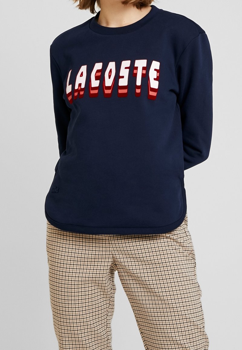 Marinblå crewneck sweatshirt i bomullsmix, med fet röd och vit "LACOSTE" text. Bärs med rutiga beige byxor.