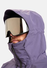 Uomo che indossa una giacca con cappuccio viola e grandi occhiali da sci riflettenti, con il volto parzialmente coperto dal colletto zip della giacca.
