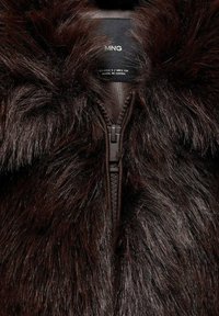 Chaqueta de piel sintética marrón oscuro con cremallera de cuero suave, que presenta un alto cuello y suave pelaje texturizado en toda la prenda. Etiqueta de tamaño visible.