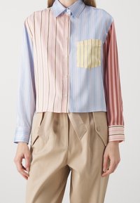Gestreepte, korte shirt met blauwe, roze en gele kleuren. Voorzien van een borstzak en knoopsluiting. Lange mouwen met contrasterende manchetten.