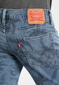 Blue denim jeans with a camouflage pattern, featuring a leather waistband label and a red tab on the back pocket.