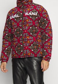 Red patterned puffer jacket featuring a geometric design with multicolored accents, zip closure, and elastic cuffs. Logo displayed on the front.