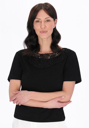 Femeie cu păr ondulat, de lungime medie, de culoare închisă, purtând un top cu guler din dantelă neagră și pantaloni albi, stând cu brațele încrucișate pe un fundal alb.