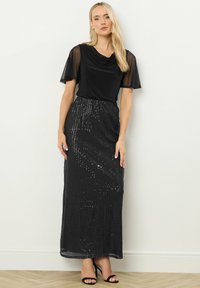 Blusa nera con scollatura drappeggiata e maniche corte in tessuto trasparente, abbinata a una gonna lunga nera con paillettes e sandali neri con tacco alto.