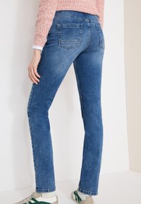 Personne portant un jean en denim bleu avec des poches arrière, un haut en tricot rose et des baskets blanches à rayures vertes, debout contre un mur blanc.
