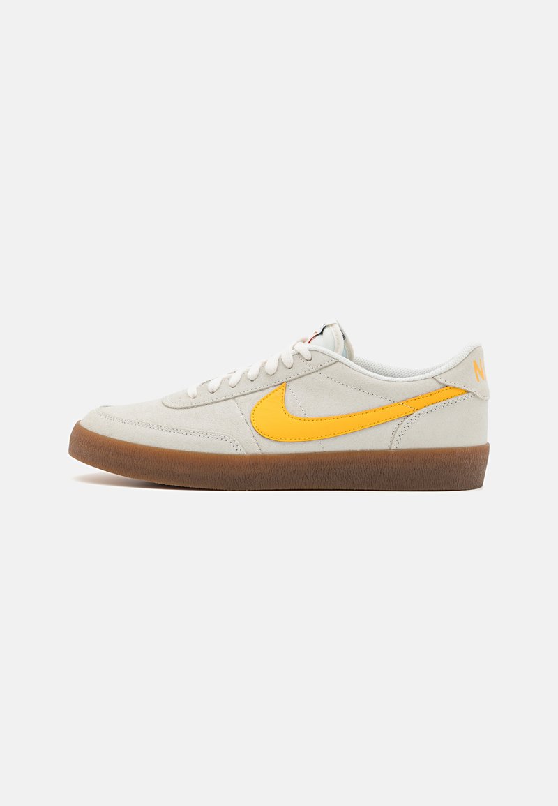 Nike tenisky ve světle šedé semišové kůži s žlutým logem Swoosh, bílými šňůrkami a hnědou gumovou podrážkou. Mají strukturovaný povrch a polstrovaný límec.