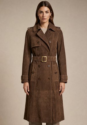 Kvinde iført en lang brun ruskind dobbeltbreddet trenchcoat med bælte og skulderpuder, stående foran en beige baggrund.