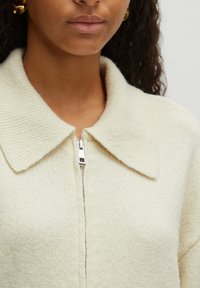 Beige Zip-Cardigan mit strukturiertem Stoff, verfügt über einen großen Kragen und einen metallischen Reißverschluss an der Vorderseite. Weiches und gemütliches Erscheinungsbild.