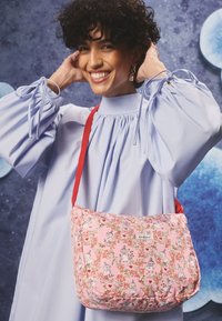 Roza torba za ramo z vzorcem, cvetličnim dizajnom in risanimi liki. Mehka tkanina, zaobljena oblika in rdeča naramnica za nošenje čez ramo.