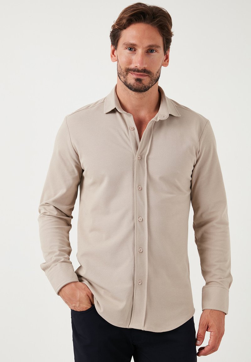Camisa de manga larga beige hecha de una tela suave y texturizada, con cuello abotonado y tapeta frontal, con botones de color marrón claro.