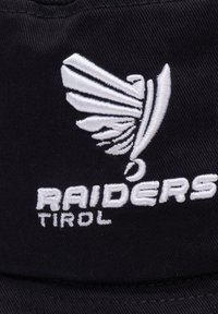 Černá čepice s bílým vyšitým logem křídla a textem "RAIDERS TIROL" níže. Design je jednoduchý s hladkým textilním povrchem.