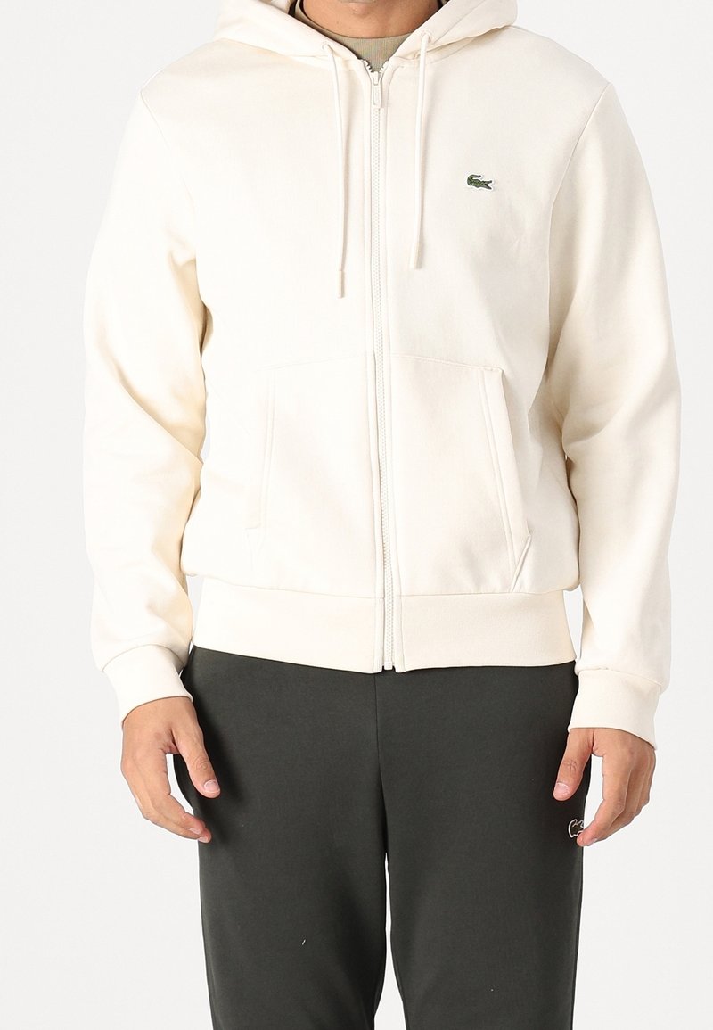 Homme portant un sweat à capuche crème zippé avec poches avant et pantalon noir, debout devant un fond blanc uni.