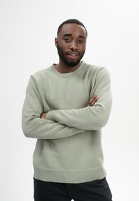 Hellgrüner gestrickter Pullover mit geripptem Saum und Bündchen. Der Stoff wirkt weich und strukturiert. Das Model trägt schwarze Hosen und schlägt die Arme übereinander.