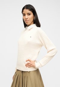 Γυναίκα που φοράει εκρού πουλόβερ με πλεξούδες πάνω από λευκό πουκάμισο με γιακά και φούστα σε σκούρο ελιάς με πιέτες, πόζαροντας με το ένα χέρι στη μέση.