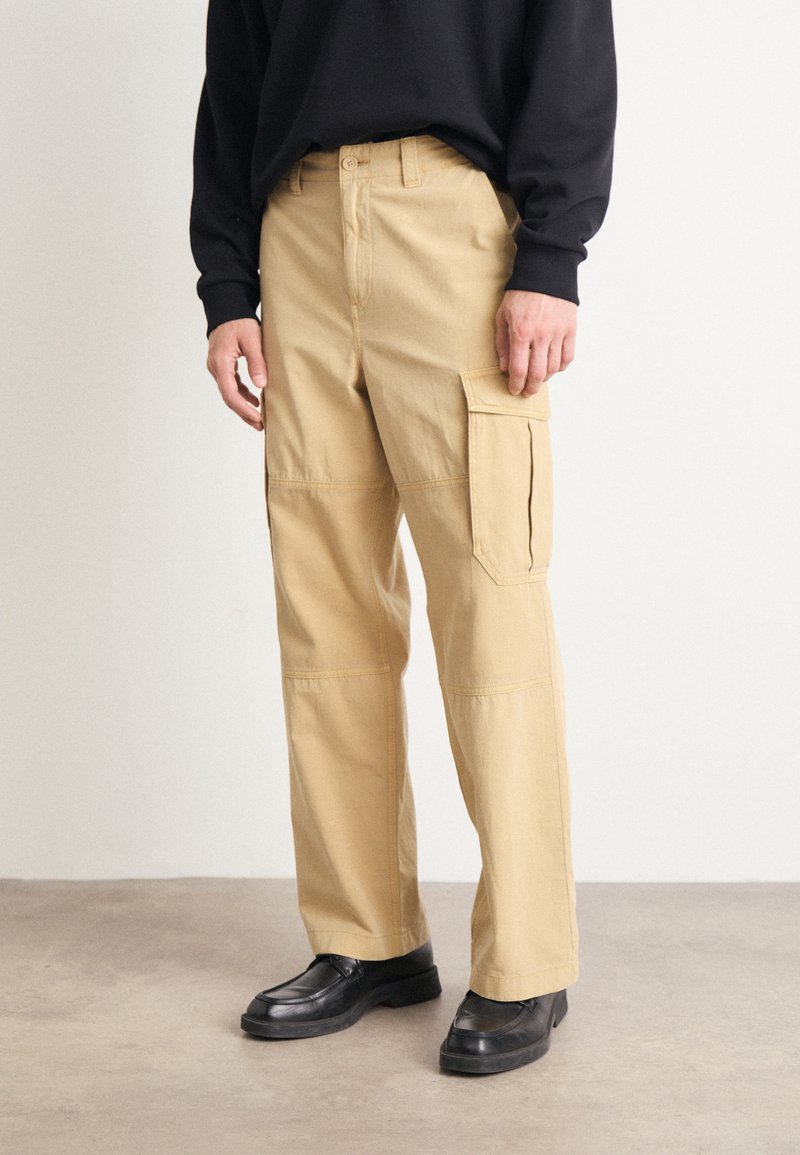 Pantaloni cargo beige con taglio dritto, dotati di tasche laterali, cuciture rinforzate e un tessuto morbido e resistente. Indossati con scarpe nere.