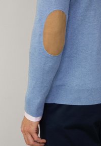 Helles blaues Strickpullover mit Ellbogenpatches aus braunem Wildleder, mit gerippten Bündchen und Saum. Schnitt und Textur sind glatt.
