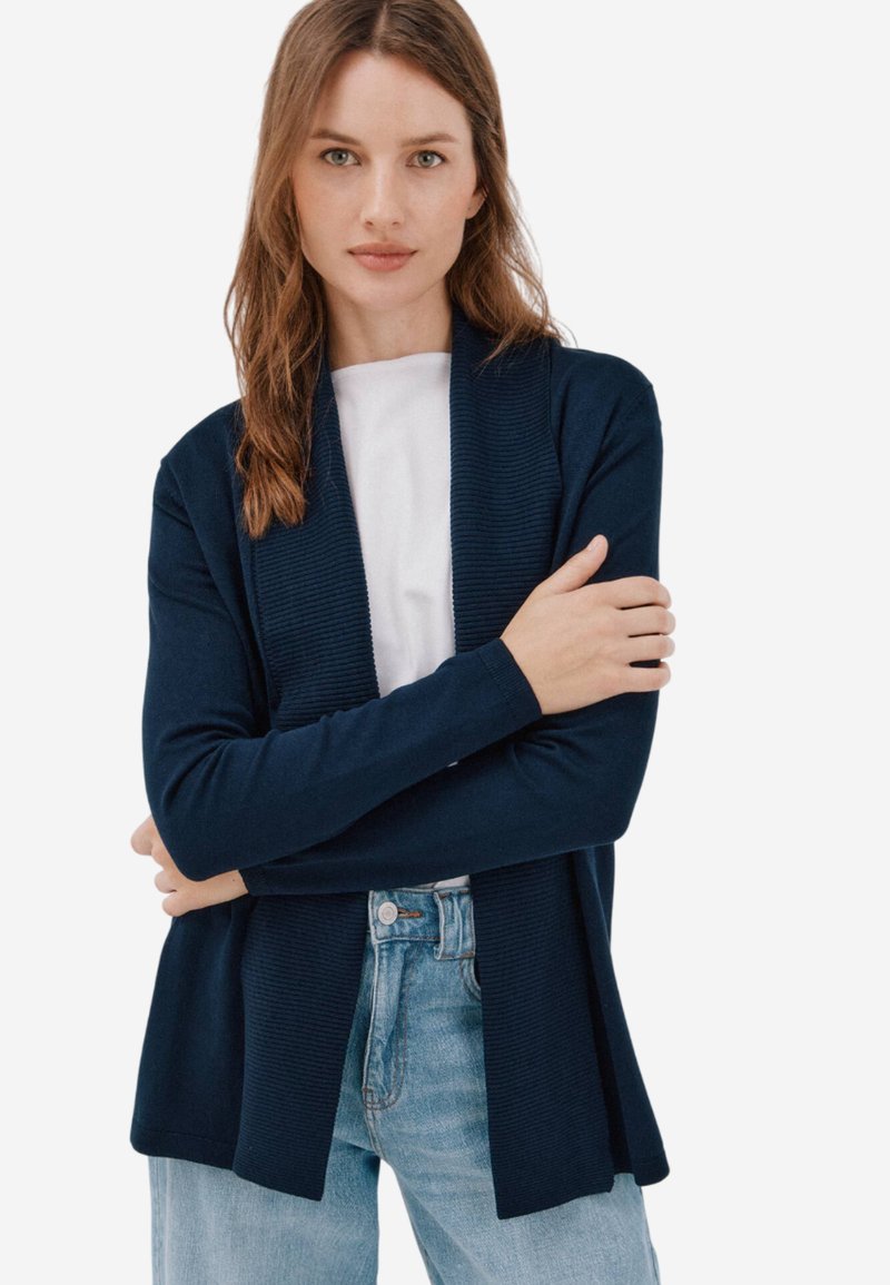 Gilet bleu marine avec texture côtelée et design ouvert à l'avant. Porté sur une chemise blanche et associé à un jean bleu clair.