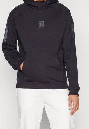 Osoba ubrana w czarną bluzę Champion z kapturem, kieszenią z przodu i logo na klatce piersiowej, w białych spodniach, stojąca z jedną ręką w kieszeni.