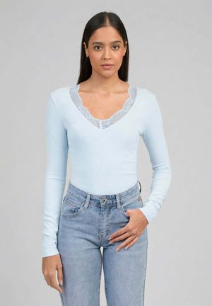 Femme aux longs cheveux bruns portant un pull bleu clair à col en V avec bordure en dentelle et un jean bleu taille haute, se tenant devant un fond gris.