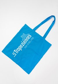 Bolsa de algodón azul con asas largas. Presenta un texto impreso en blanco que dice "Tropeziennes par M. Belarbi" y un gráfico estilizado arriba.