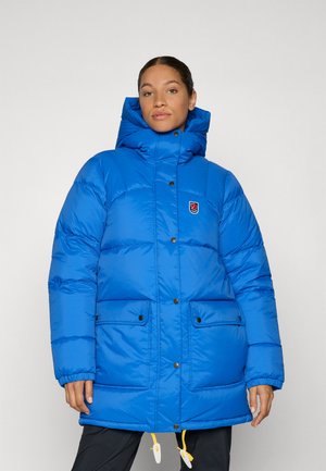 Vrouw in een felblauwe gewatteerde winterjas met een hoge kraag, drukknoopjes, twee voorzakken met flap en gele koordjes aan de zoom.
