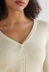 Jasbeżowy kardigan o prążkowanej fakturze z głębokim dekoltem w serek i pojedynczym zapięciem na guzik z przodu. Miękki, przytulny materiał z delikatnym połyskiem.