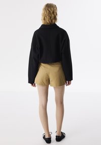 Schwarze, cropped Jacke mit weiten Ärmeln, kombiniert mit sandfarbenen Shorts mit ausgefransten Kanten. Schwarze Schnürschuhe runden den Look ab.
