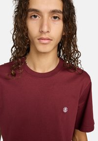 Bordeaux-farbenes Baumwoll-T-Shirt mit Rundhalsausschnitt und kurzen Ärmeln. Verfügt über ein kleines gesticktes Logo auf der linken Brustseite. Glatte Textur.