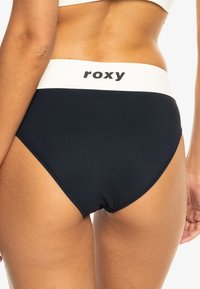 Marineblauwe bikini onderkant met een witte elastische tailleband met de merknaam "Roxy" in het zwart. Glad materiaal met een klassieke snit.