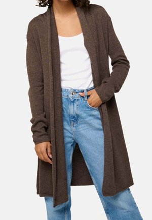 Frau trägt eine lange braune Strickjacke mit offenem Vorderteil über einem weißen Tanktop und hellblauen, hoch geschnittenen Jeans, eine Hand in der Tasche.