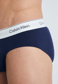 Dunkelblaue Baumwollslips mit einem weißen elastischen Bündchen, das das Logo "Calvin Klein" trägt. Glatte Textur und taillierte Passform.