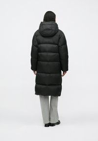 Pessoa com cabelo cacheado a usar um longo casaco acolchoado preto com capuz, calças cinzentas claras e sapatos pretos, de pé, virada de costas, em fundo branco.