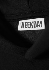 Sort stoff med en myk tekstur, med en hvit etikett som har påskriften "WEEKDAY" i fete, store bokstaver.