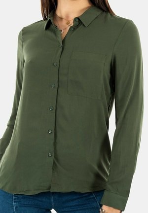 Femme portant un chemisier boutonné vert olive avec une poche sur le devant, associé à un jean bleu et des colliers en or superposés.