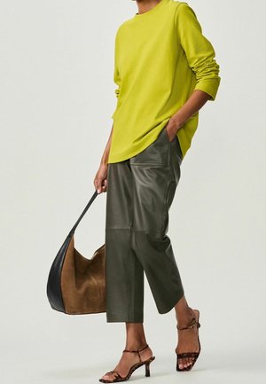 Osoba w jaskrawozielonej, długorękawowej koszuli, krótkich skórzanych spodniach w ciemnozielonym kolorze, brązowych sandałach na obcasie w panterkę, trzymająca brązową i czarną torbę na ramię.