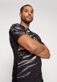 Fekete sportpóló, amely texturált mintával és fehér grafikus vonalakkal rendelkezik. Rövid ujjak és kerek nyakkivágás jellemzi. Az anyag simának tűnik.