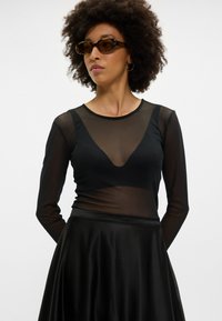 Fekete, hosszú ujjú blúz áttetsző anyagból, mély V-kivágással, sima fekete selyemszoknyával párosítva. Teknősbéka mintázatú napszemüveggel kiegészítve.